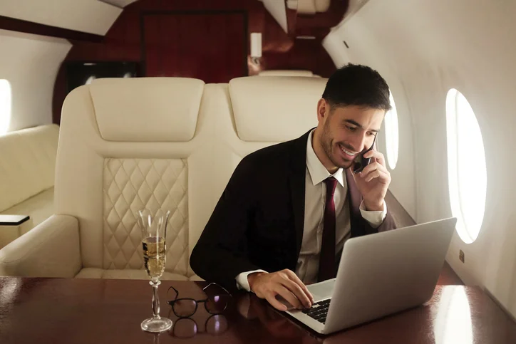 man-in-a-suit-smiling-while-talking-on-the-phone-and-working-on-a-laptop-inside-a-private-jet